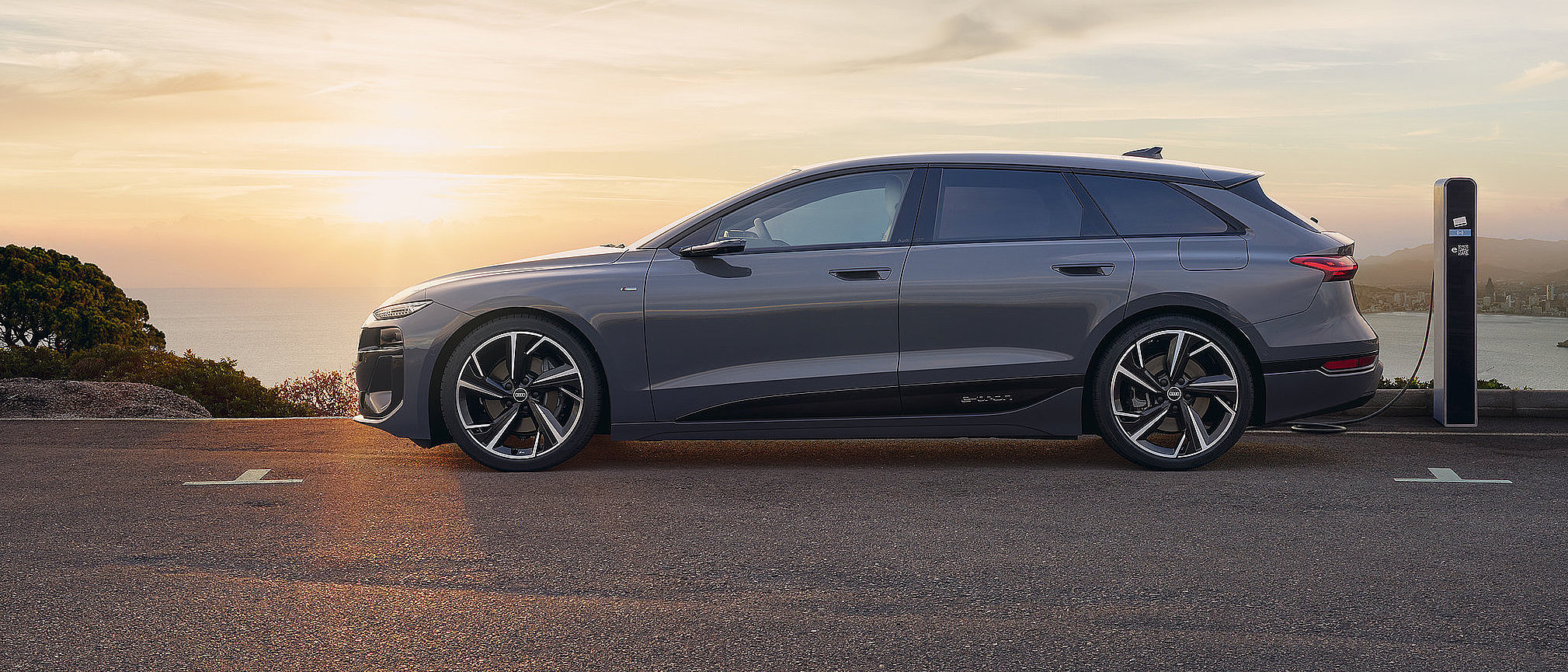 Grauer Audi A6 Avant e-tron lädt an einer Ladesäule bei Sonnenuntergang mit Blick auf die Küste.