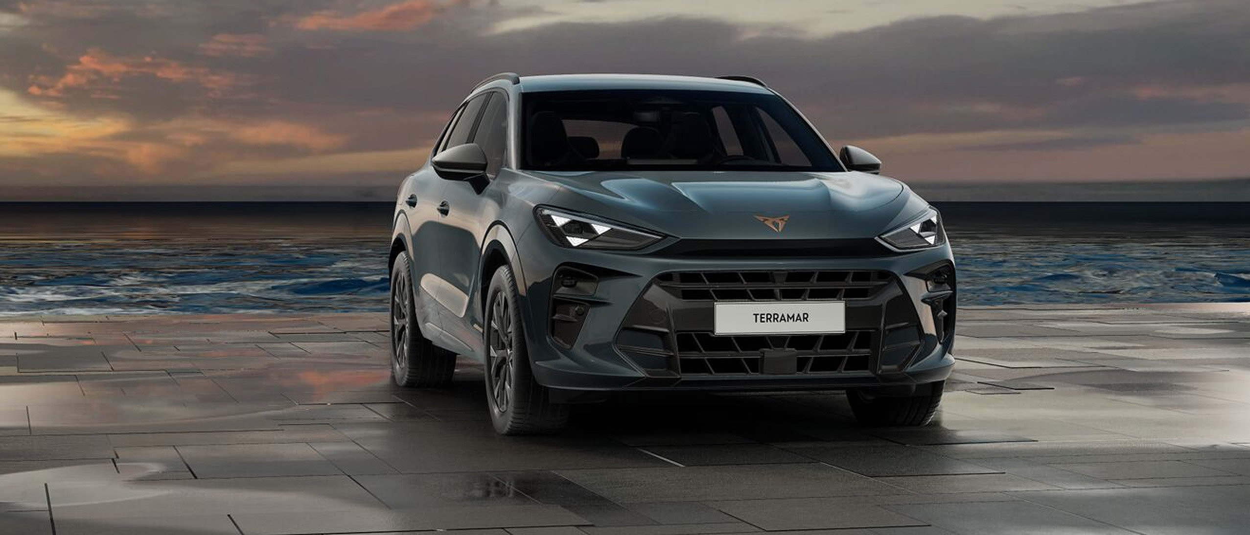 CUPRA Terramar in Fiord Blau in Frontansicht auf nasser Küstenstraße vor Sonnenuntergang.