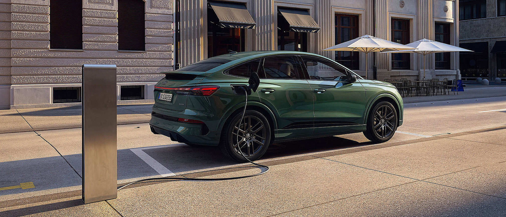 Grüner Audi Q4 e-tron Elektro-SUV beim Laden an einer öffentlichen Ladestation vor einem modernen Stadtgebäude.