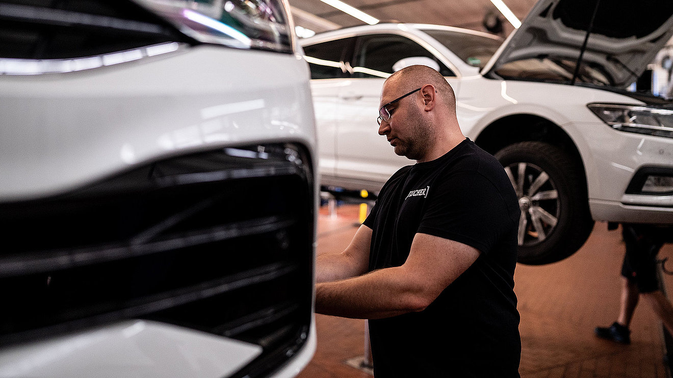 Werkstattmitarbeiter führt an einem weißen Fahrzeug auf einer Hebebühne in der Autowerkstatt eine UVV-Prüfung durch am Rad vorn rechts und prüft Reifen und Bremse
