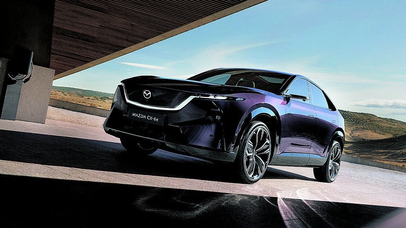 Violetter Mazda CX-6e Elektro-SUV in dynamischer Front-Seitenansicht unter moderner Architektur mit weitläufiger Landschaft im Hintergrund.