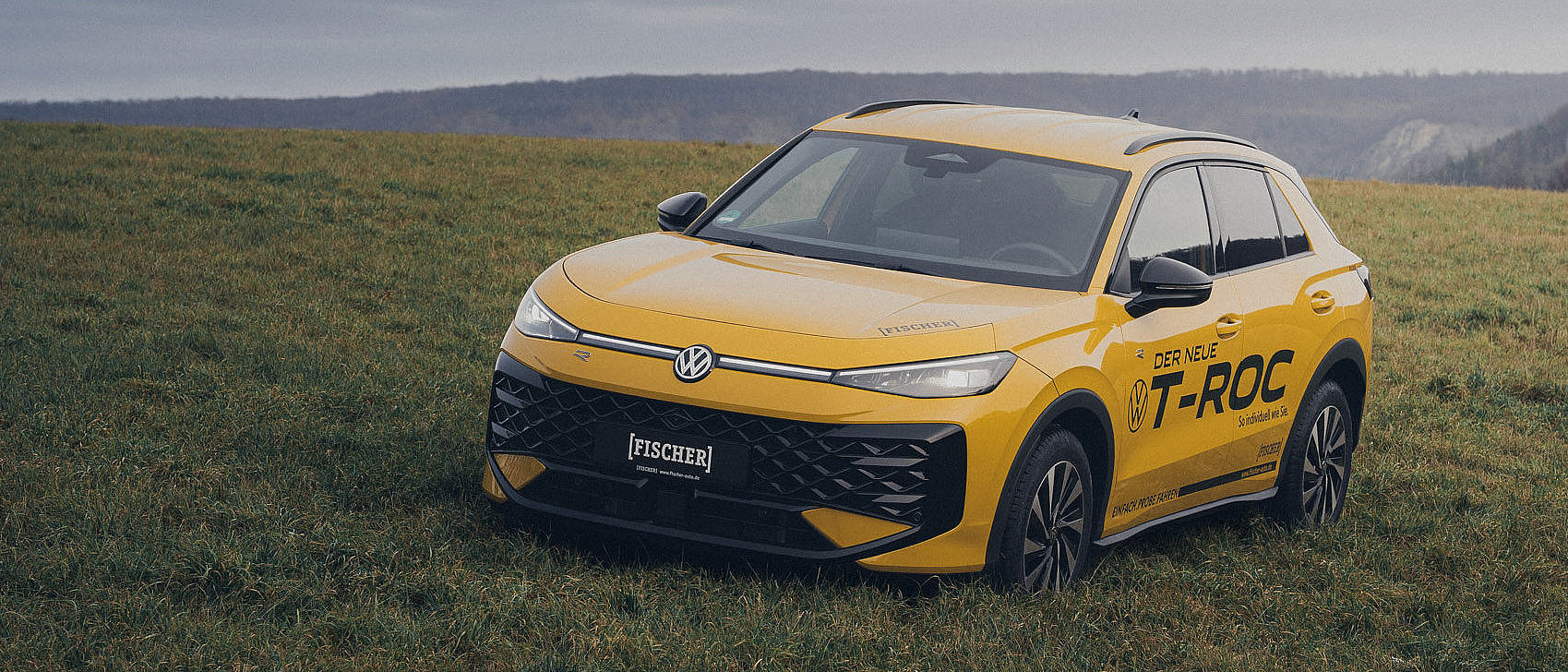 T-Roc 2026 Comercial Gelber Volkswagen T-Roc 2026 von Fischer auf einer Wiese vor hügeliger Landschaft