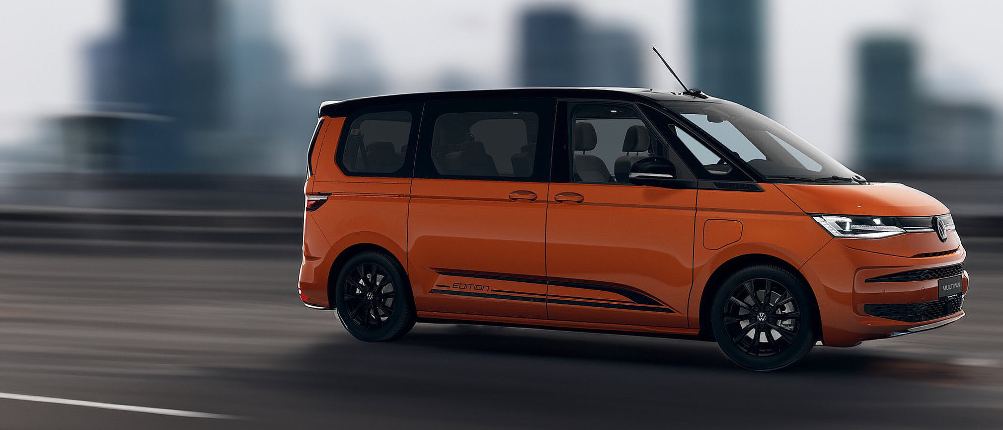 Oranger Volkswagen Multivan eHybrid Edition in Seitenansicht auf der Straße vor urbaner Kulisse.
